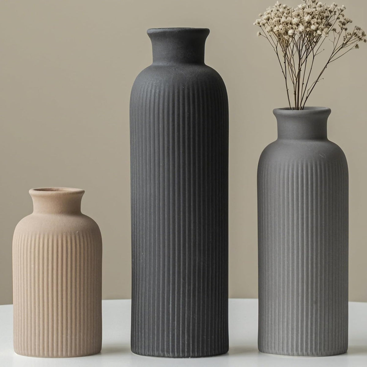Three ribbed matte ceramic vases in ascending heights—beige, tall charcoal, and mid-gray—on a white surface; dried stems in