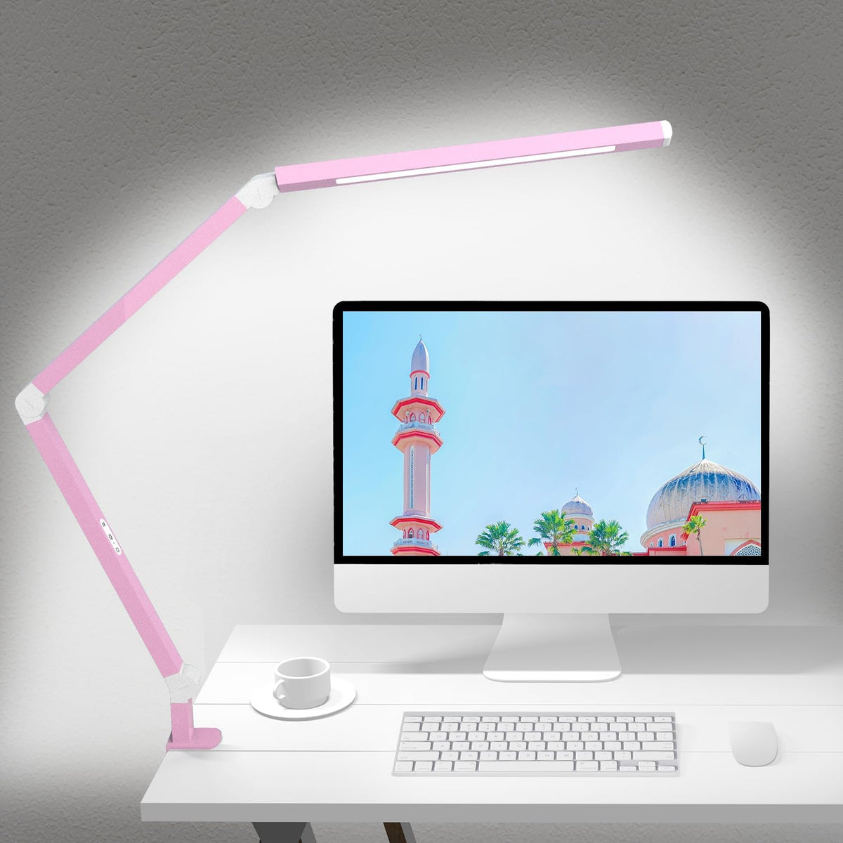 Pink dual-arm clamp lamp lit above a desktop monitor, showing the pink finish and clamp base on a white desk.