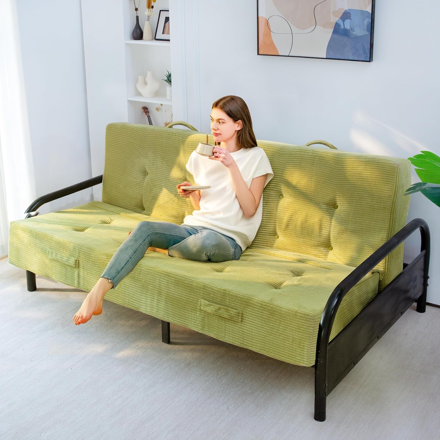 Woman sitting on a green corded futon mattress on a black metal frame, holding a cup and saucer.