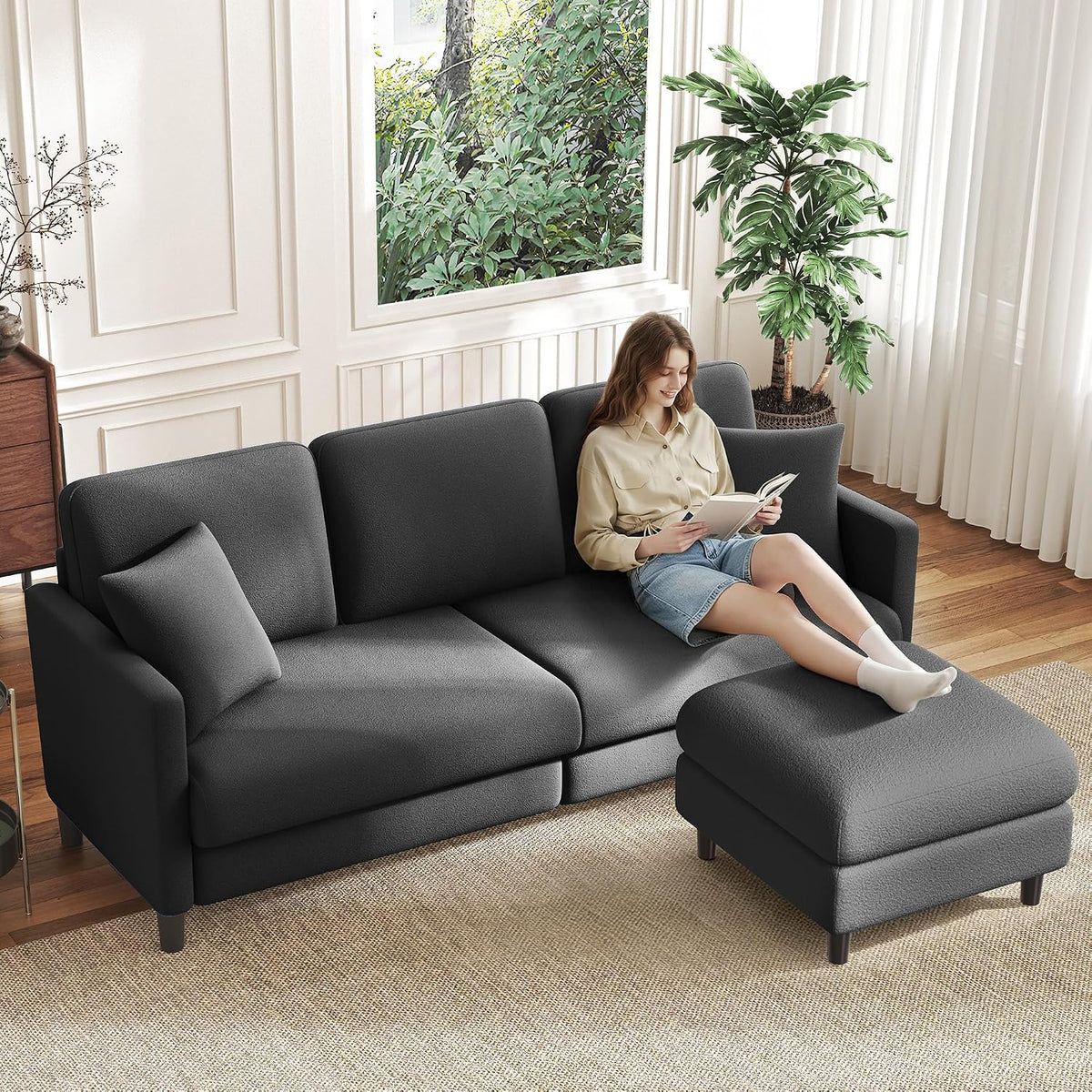 Dark gray sofa and ottoman with a woman reading, styled beside plants and a window