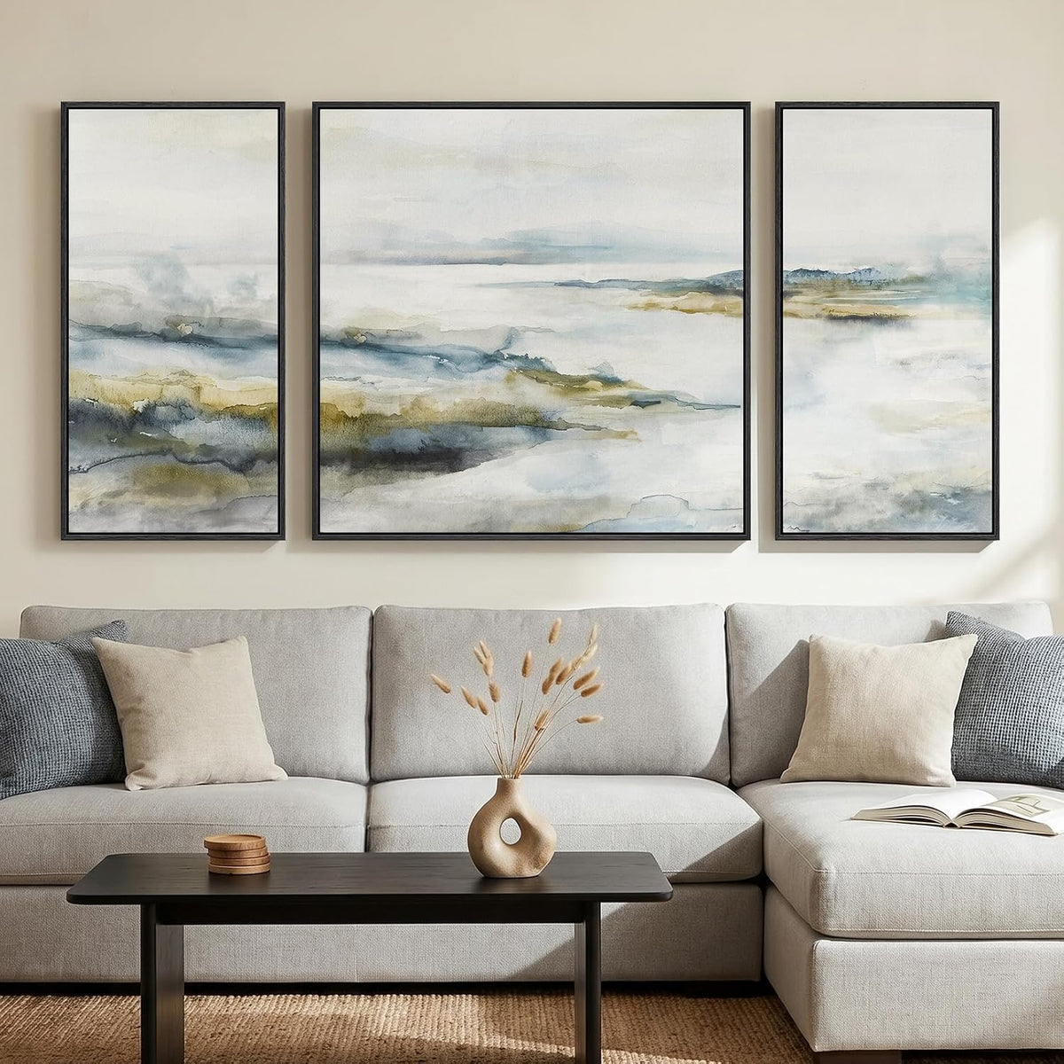 Three-panel soft watercolor landscape in pale blues and tans above a light gray sofa with a vase on the coffee table.