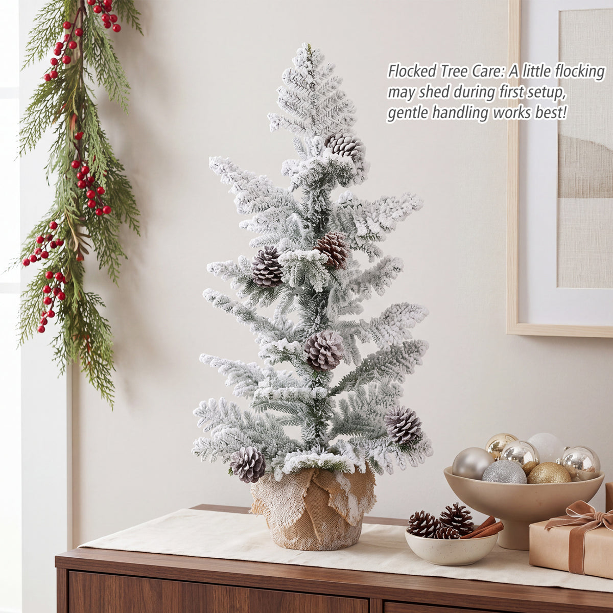 Snow-flocked tabletop tree with pinecones in a burlap-wrapped base on a cabinet.