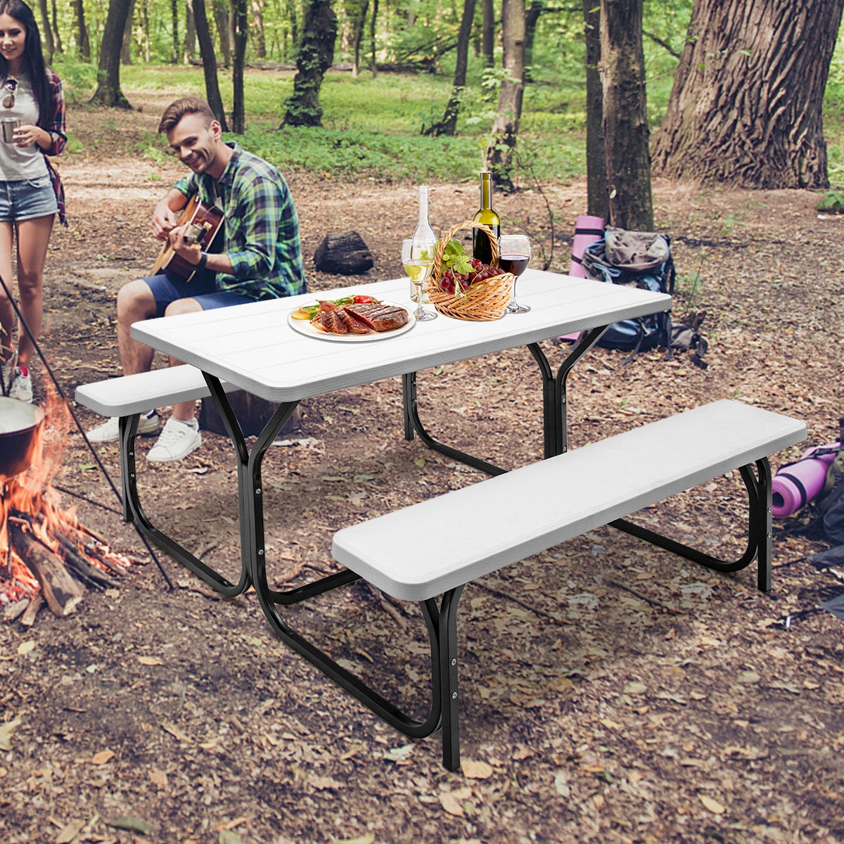 Picnic table and benches set near a campfire, with people, food, and camping gear in a wooded clearing.