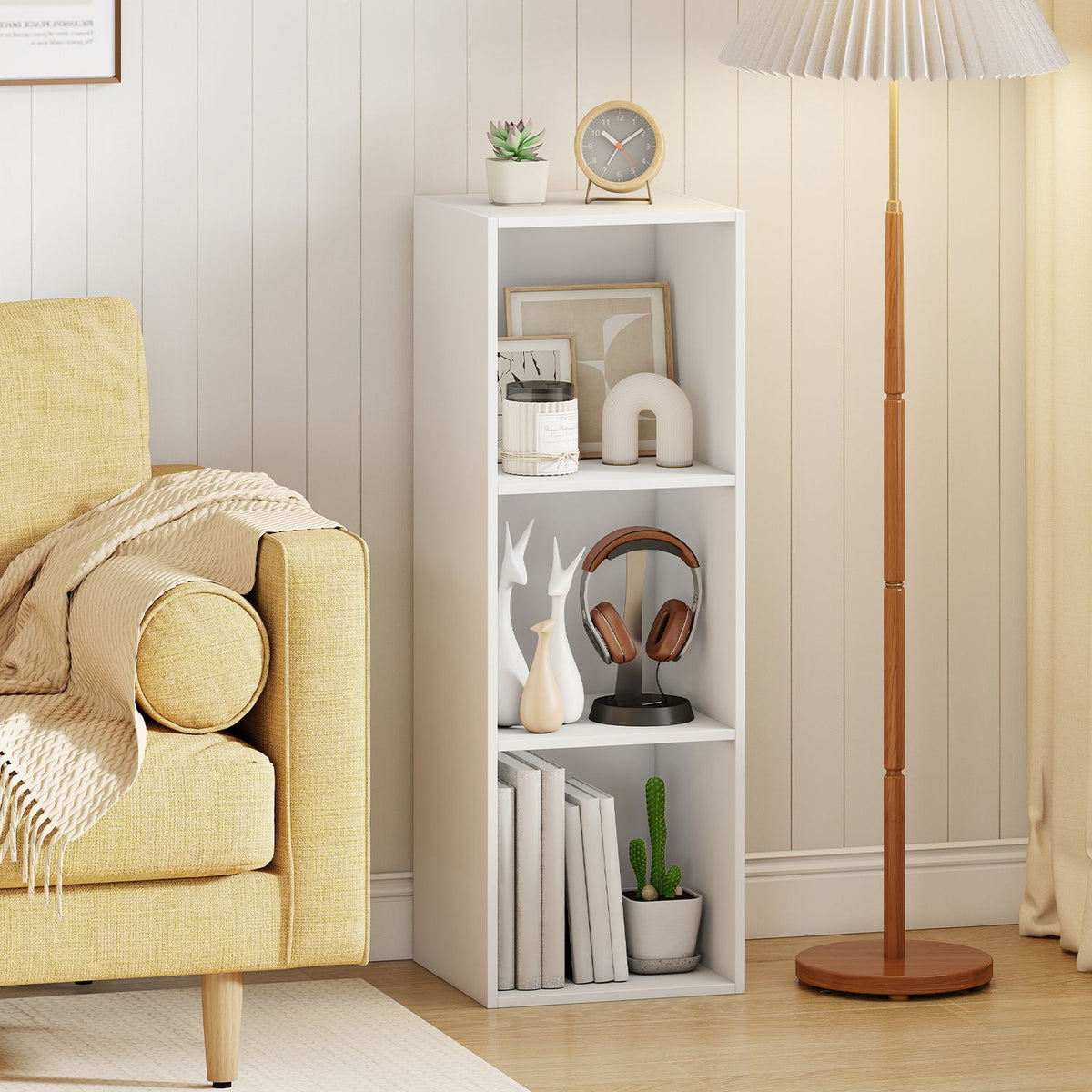 White three-cube bookshelf styled with books and decor between a yellow armchair and a wooden floor lamp.