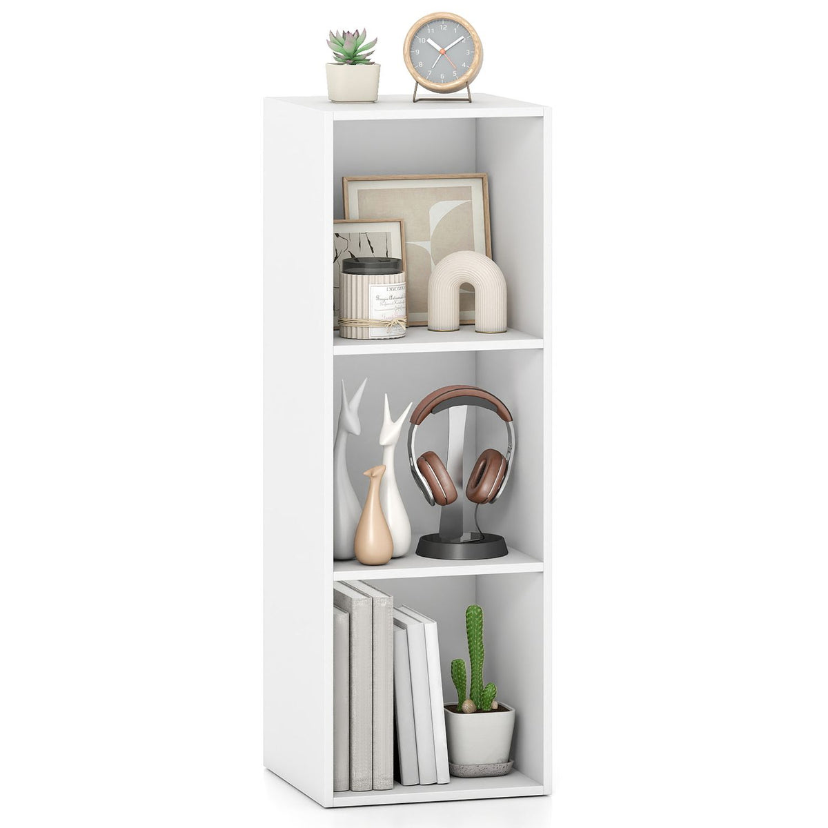 White three-cube bookshelf holding books, small ornaments and headphones, with a plant and clock on top.
