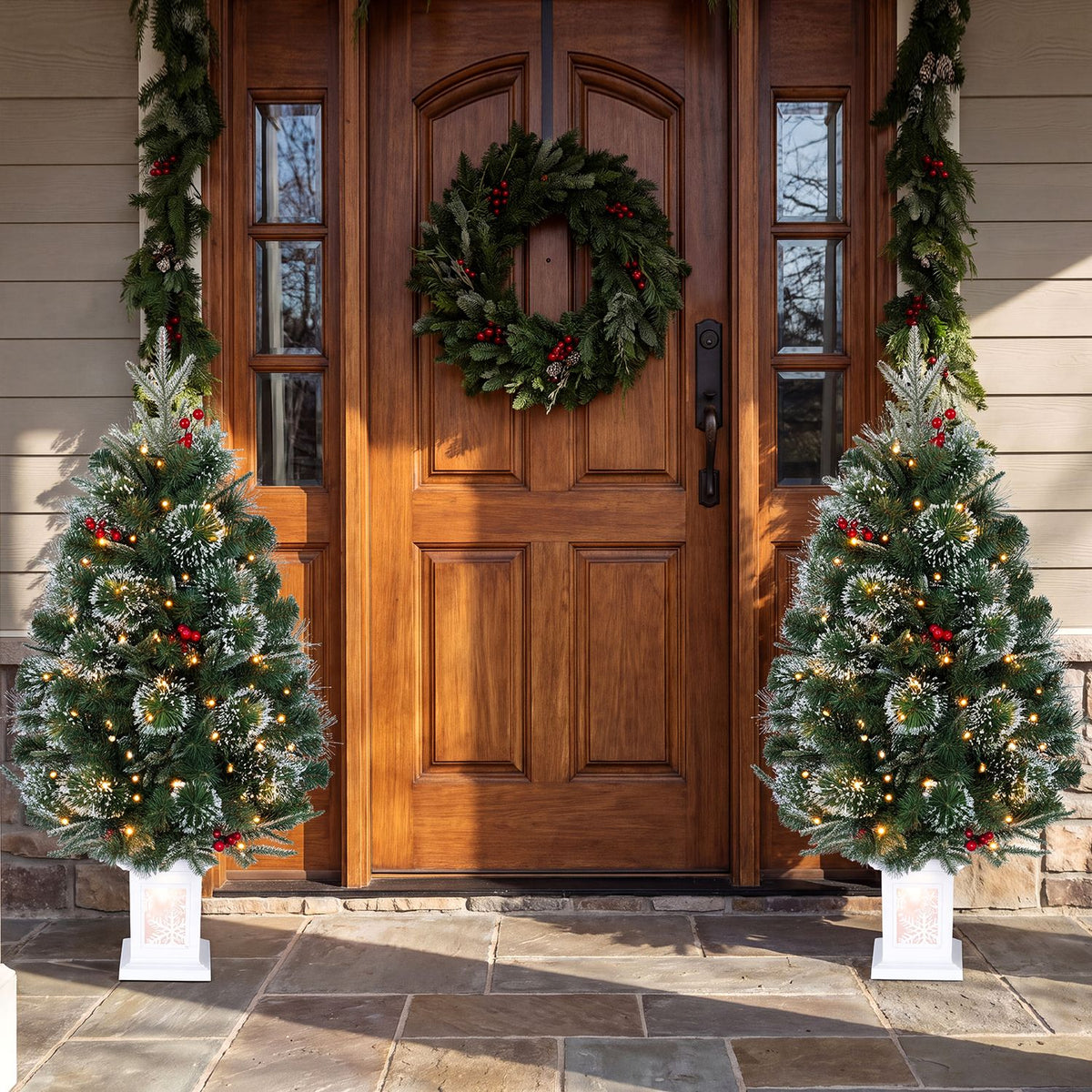 Two lit, decorated potted Christmas trees flank a wooden front door.