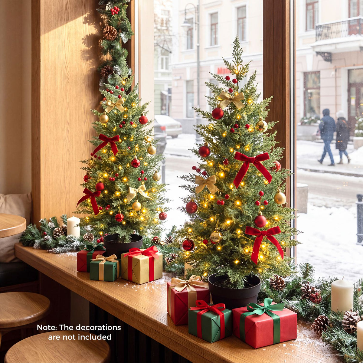 Two glowing, ornamented potted trees sit on a table by a window, with wrapped gifts around them.