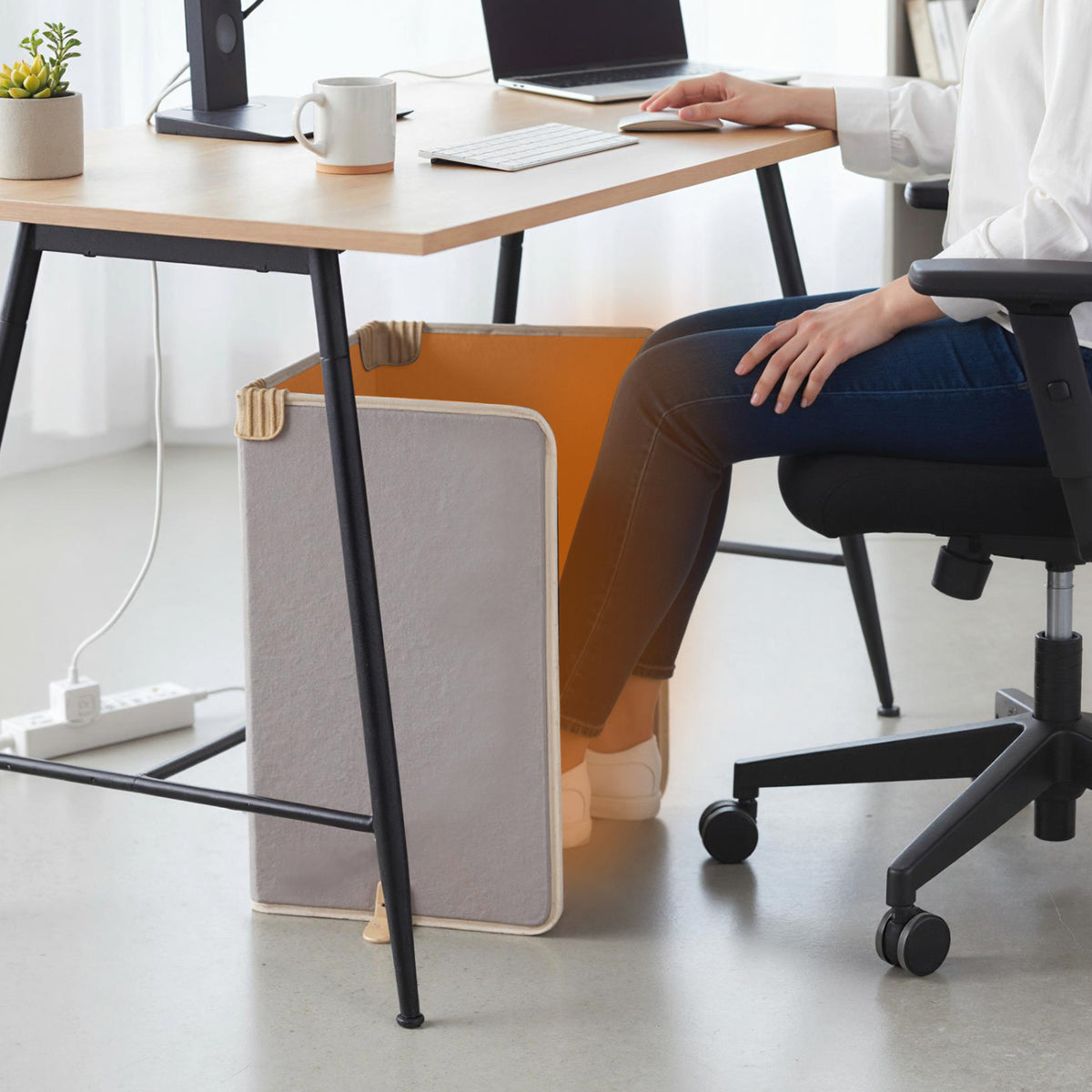 A seated person warms their legs beside a three-panel heater under a desk.