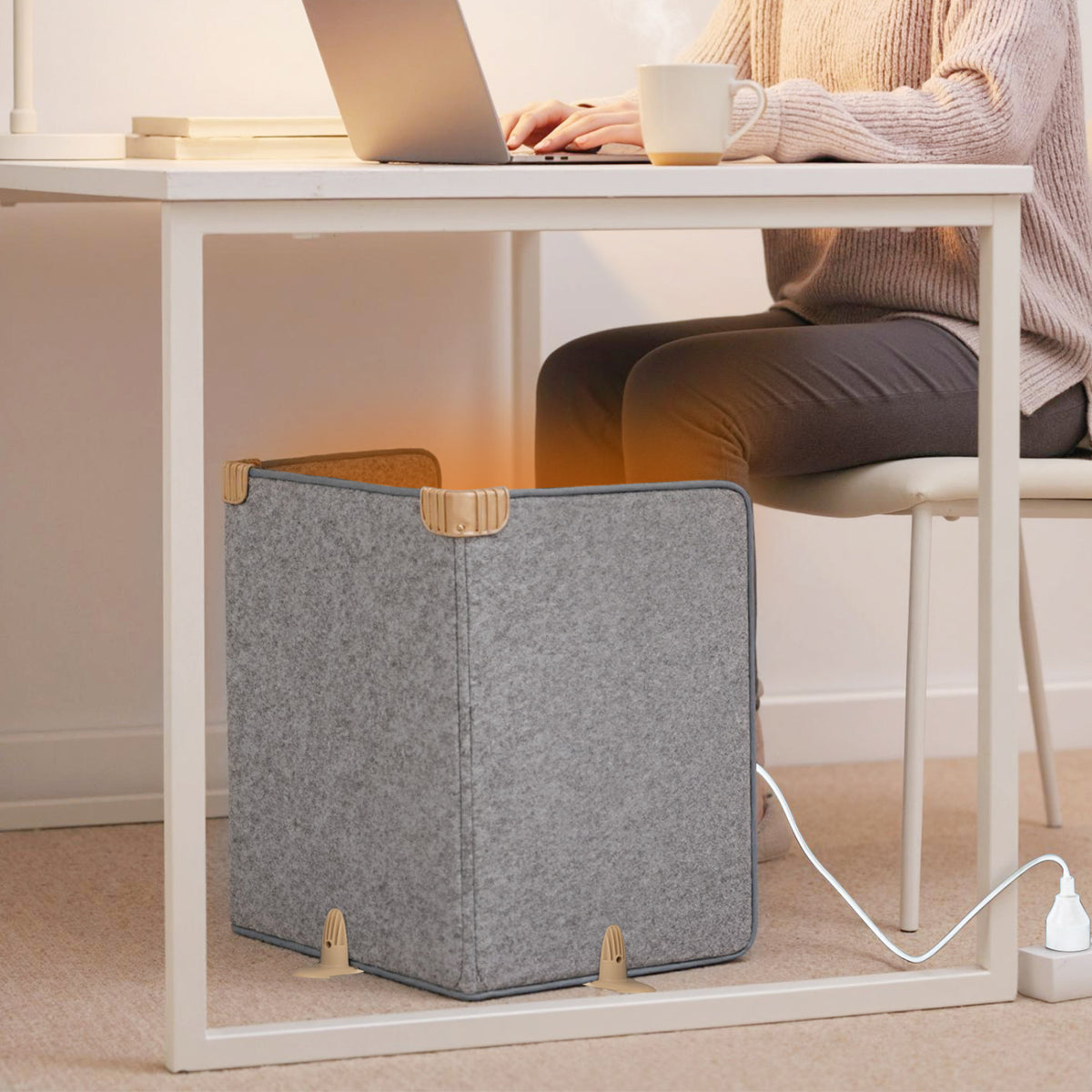 Gray folding panel heater sits beneath a desk beside a seated person, with its power cord plugged into an outlet.