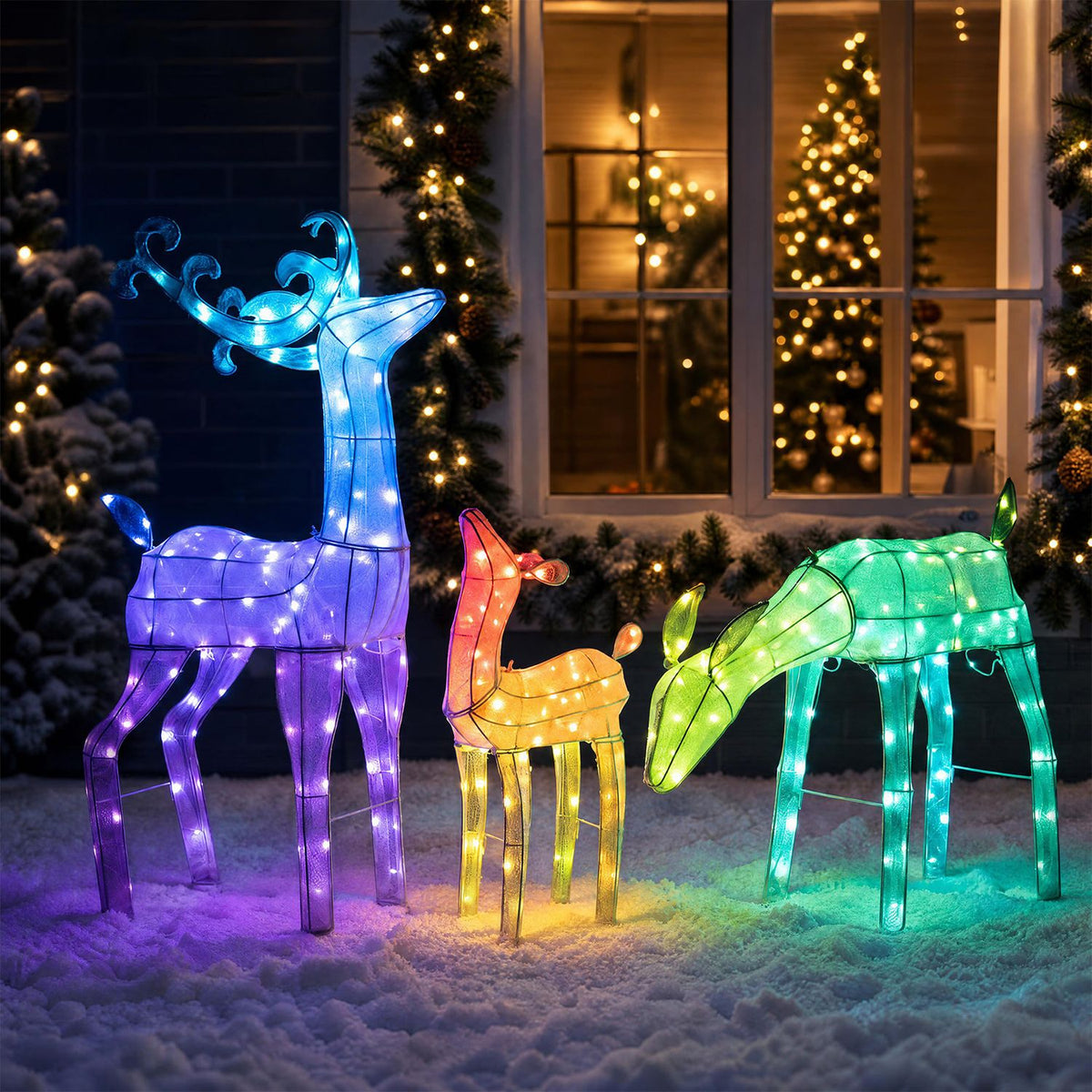 RGB-lit deer family arranged in snow outside a warmly lit house