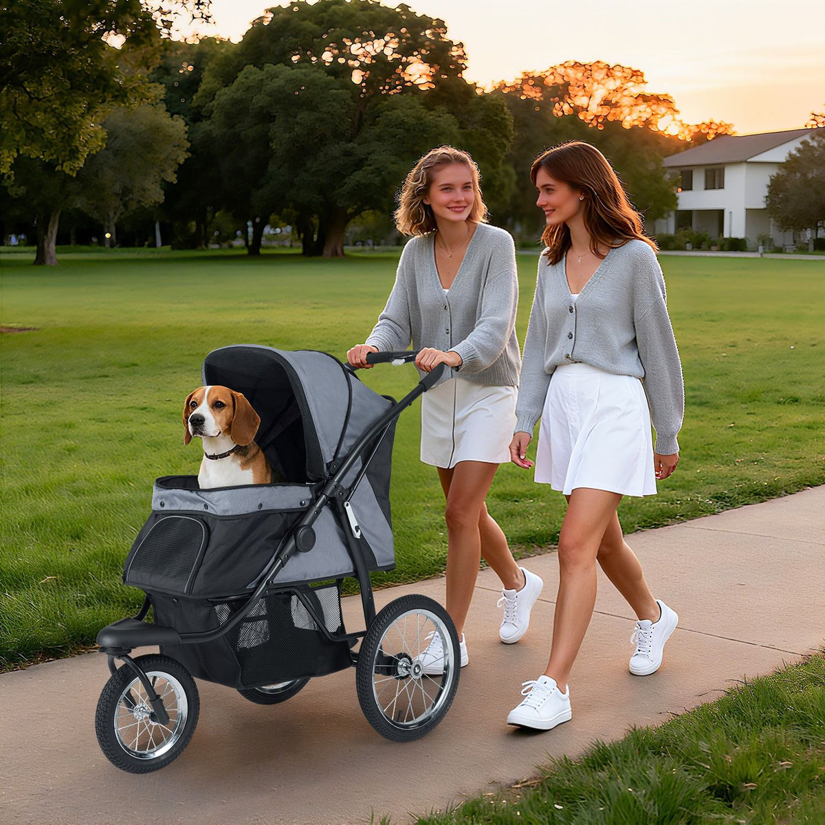 Two women walk beside a beagle riding in a gray and black pet stroller on a park path.