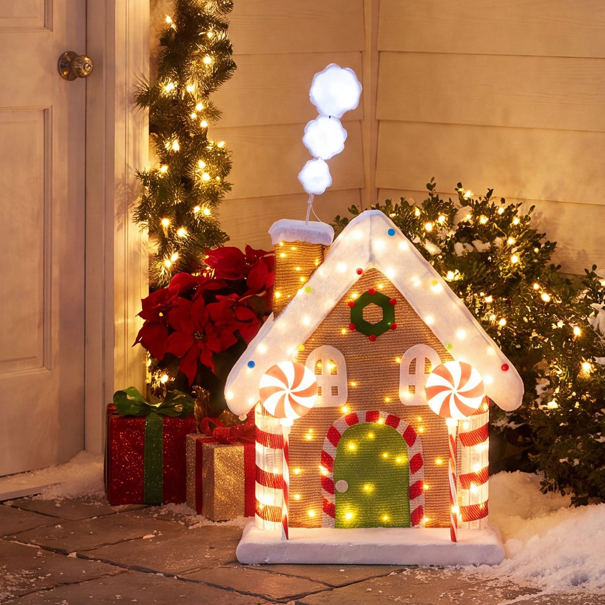 Lit gingerbread house displayed on a snowy porch beside a Christmas tree and wrapped gifts.