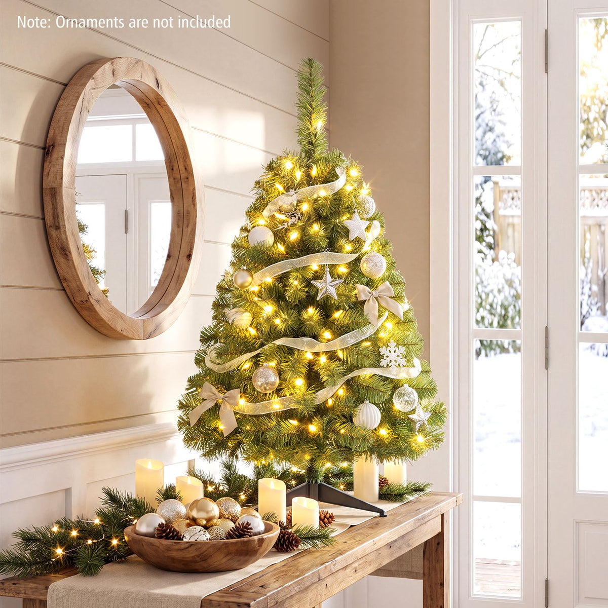 Decorated, glowing mini Christmas tree displayed on a wooden console beside a round mirror and tall windows.