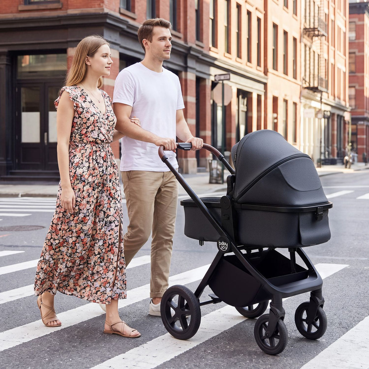 A couple walks beside a dark stroller with a covered bassinet on a city crosswalk.
