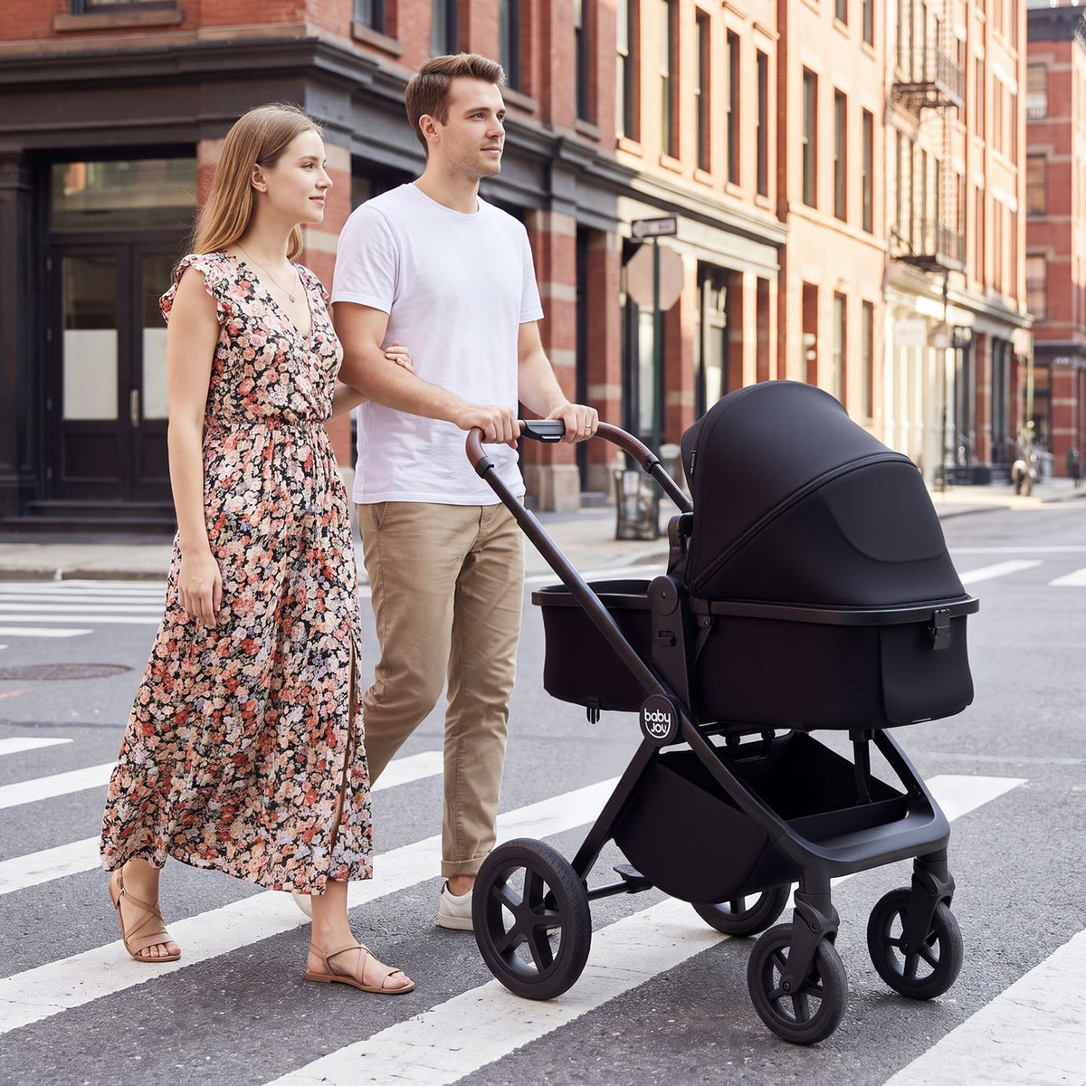A couple walks beside a black stroller with a covered bassinet on a city crosswalk.