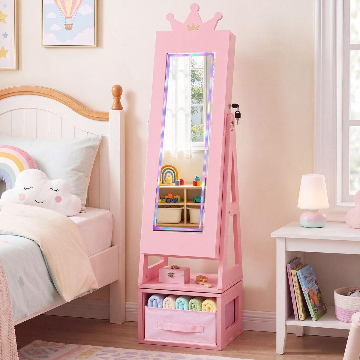 Pink crown-topped jewelry cabinet with a lit full-length mirror standing beside a bed in a child's room.