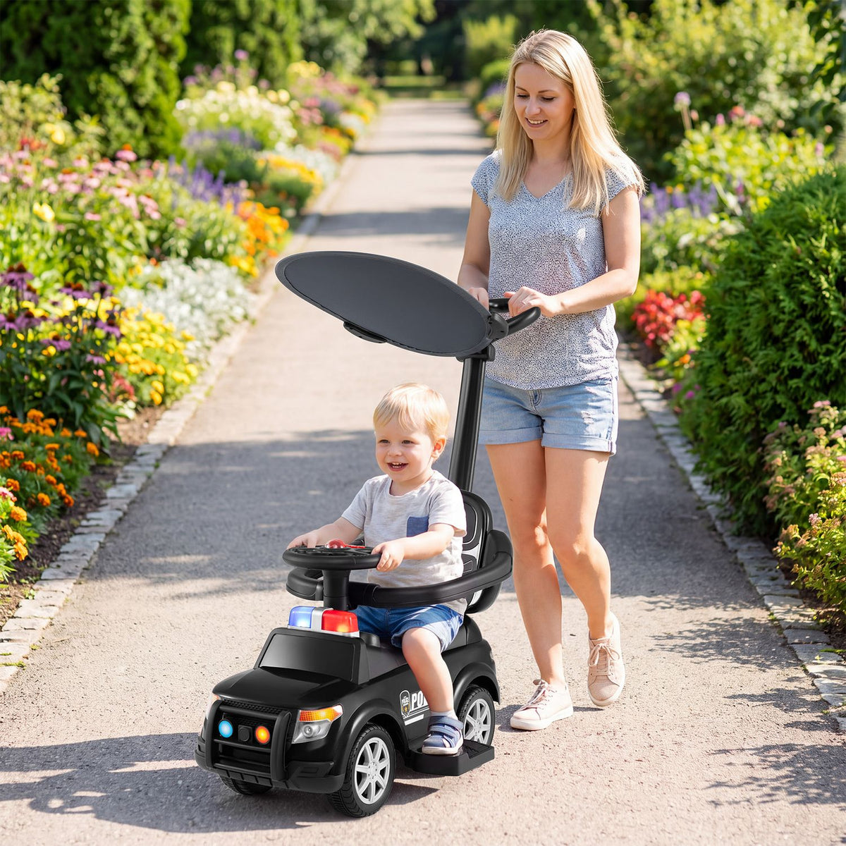 A toddler rides a black police car beneath its canopy as an adult guides it along a garden path.