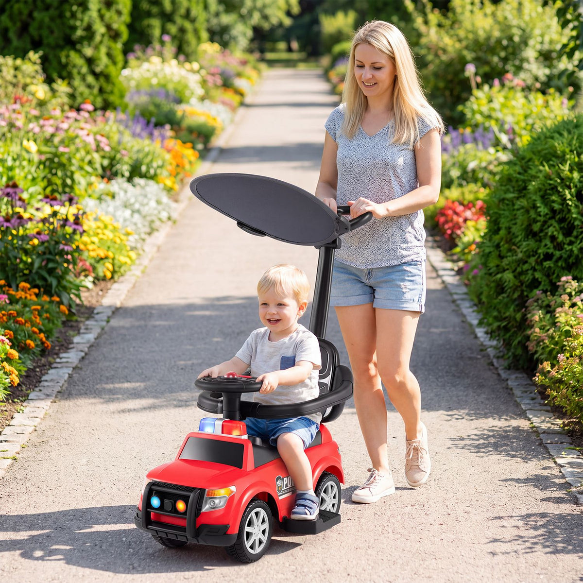 A child rides a red police car with a canopy as an adult pushes it along a garden path.