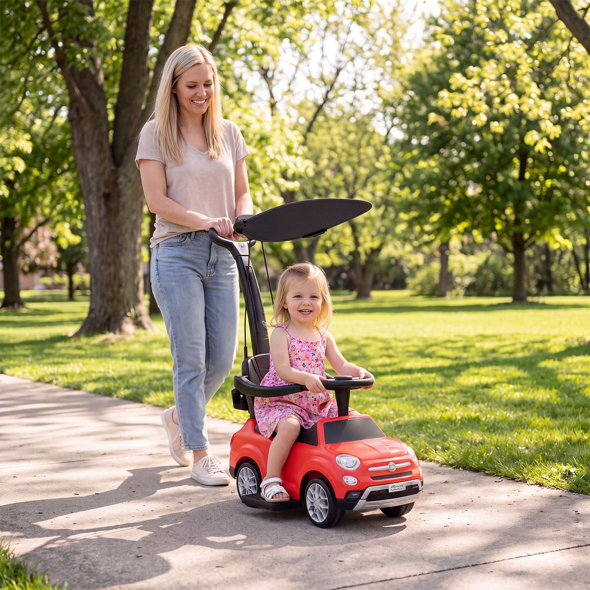 A woman pushes a smiling toddler in a red ride-on car along a sunny park path.