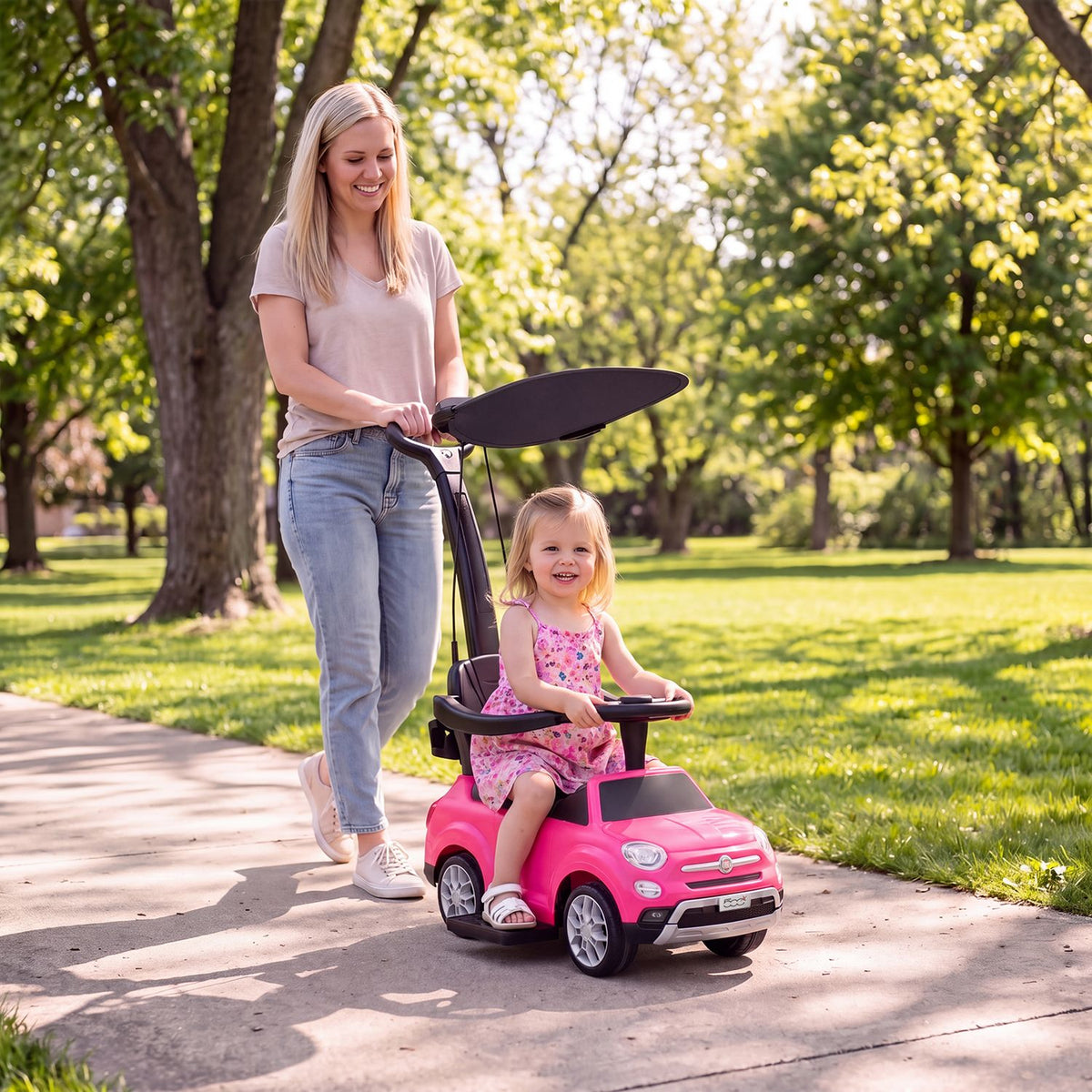 A woman pushes a smiling toddler in a pink ride-on car along a sunny park path.
