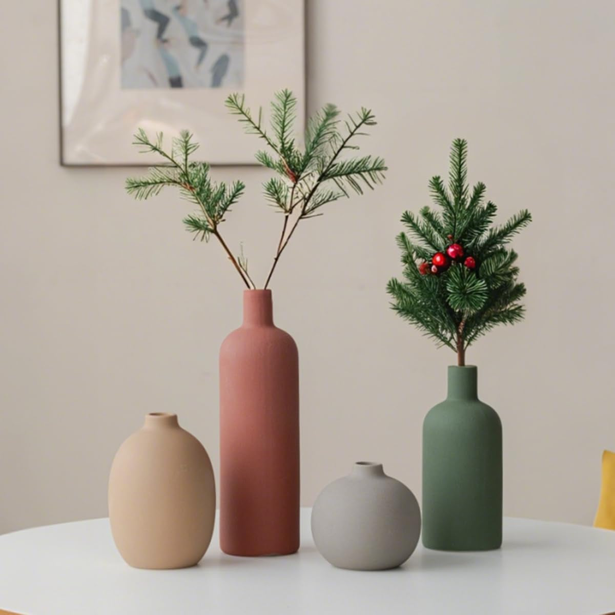 Four matte ceramic vases on a white table with pine sprigs and holiday greenery, three-quarter view