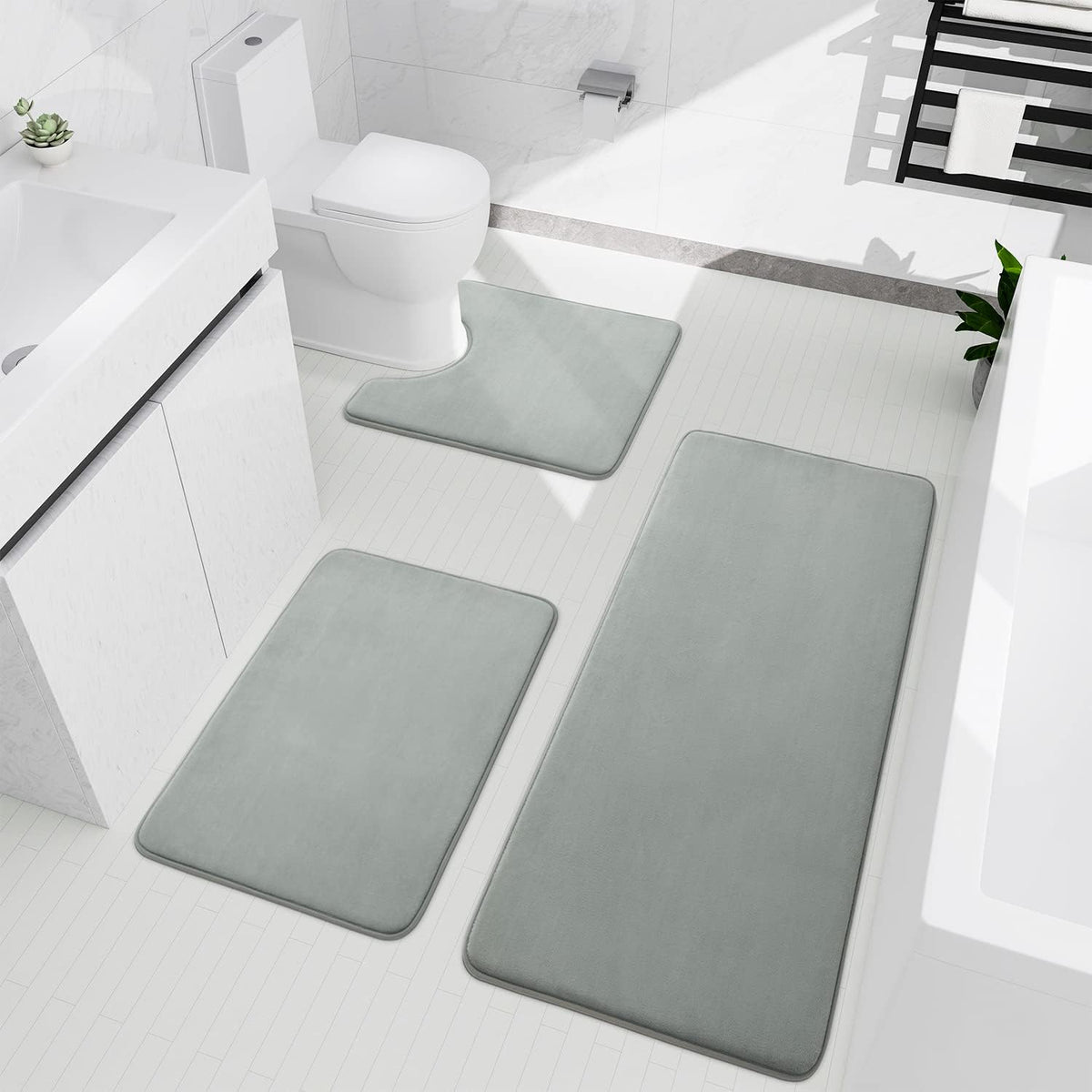 Bright bathroom seen from above showing a three-piece light gray memory-foam rug set around sink, toilet, and tub.