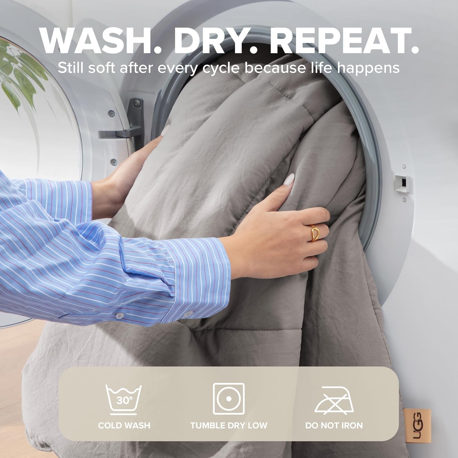 Hands placing the light grey comforter into a front‑loading dryer, showing fabric being loaded.
