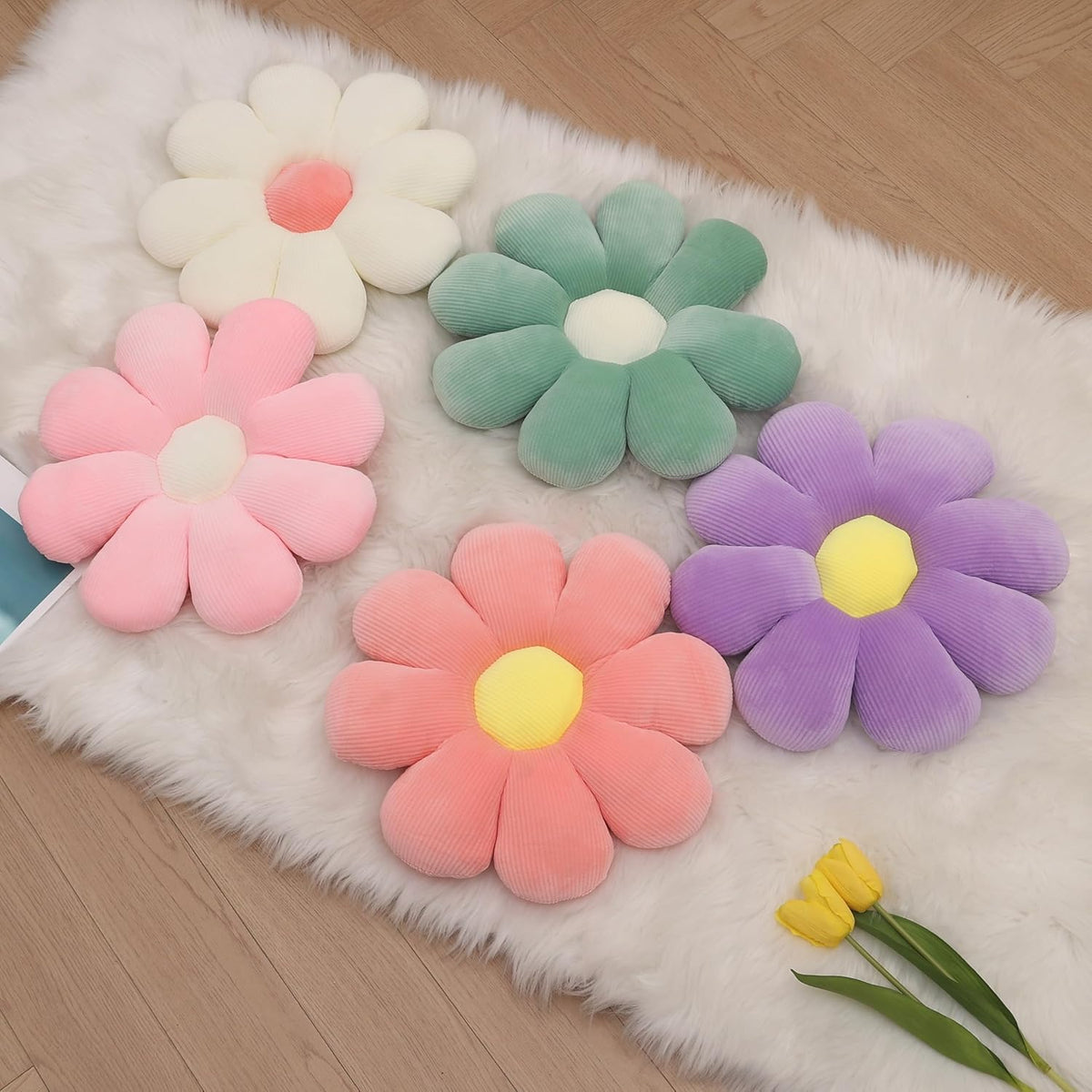 Four pastel daisy floor cushions in cream, green, pink and purple on a white faux-fur rug, top-down view