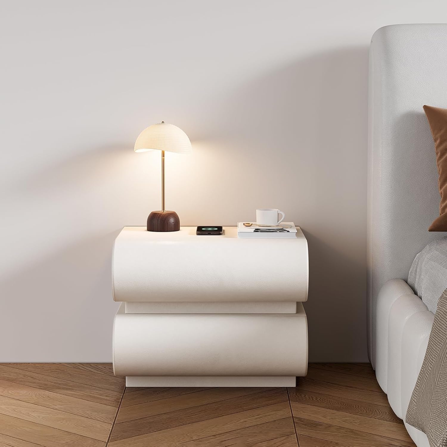 Cream-colored curved two-drawer nightstand beside a bed with lamp and cup on top