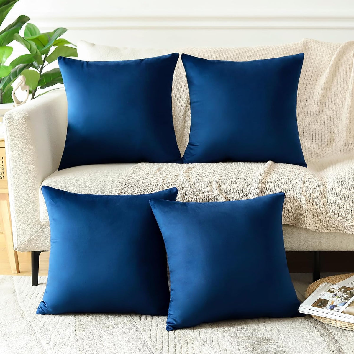 Four square navy blue velvet pillows arranged on a cream sofa with a textured throw and plant in background.