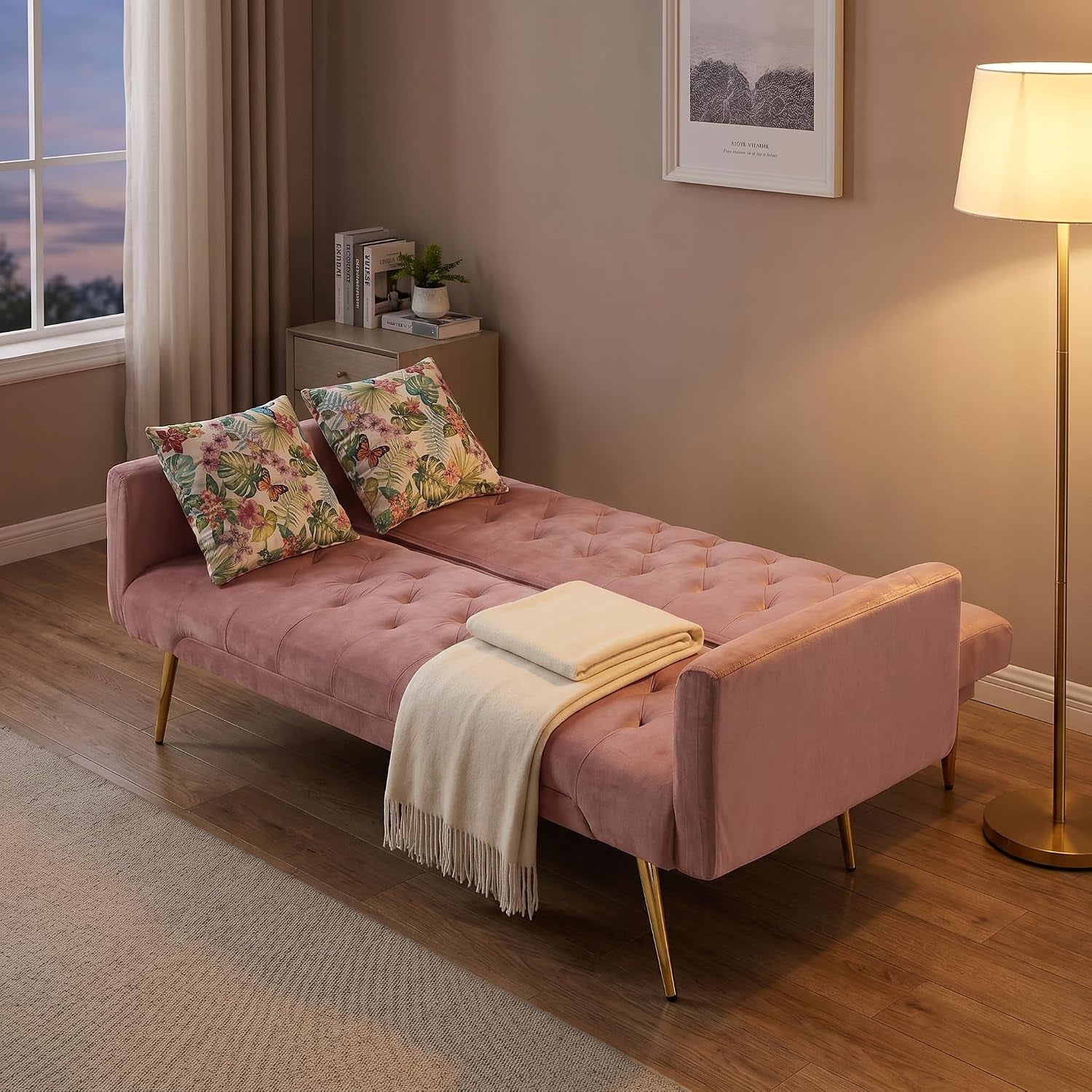 Pink velvet futon fully reclined as a bed with two floral throw pillows and a cream throw.