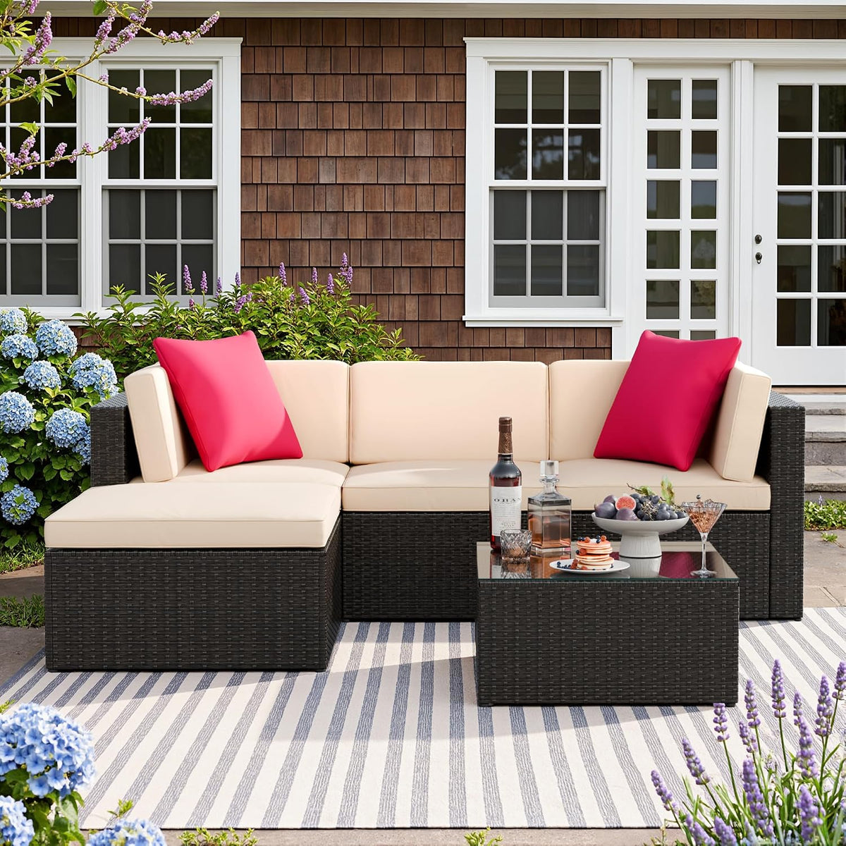 Beige cushioned wicker sectional on a striped rug outside a house with red accent pillows and a glass-top table