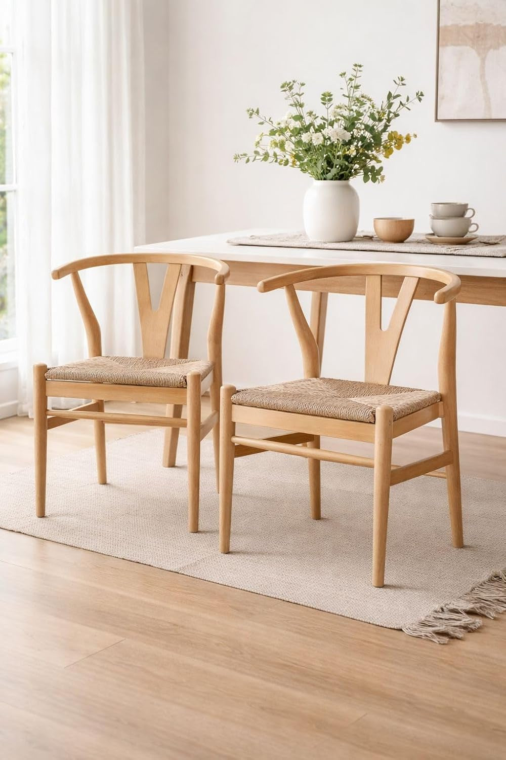 Two natural wood wishbone dining chairs beside a wood table on a neutral rug in a bright dining room