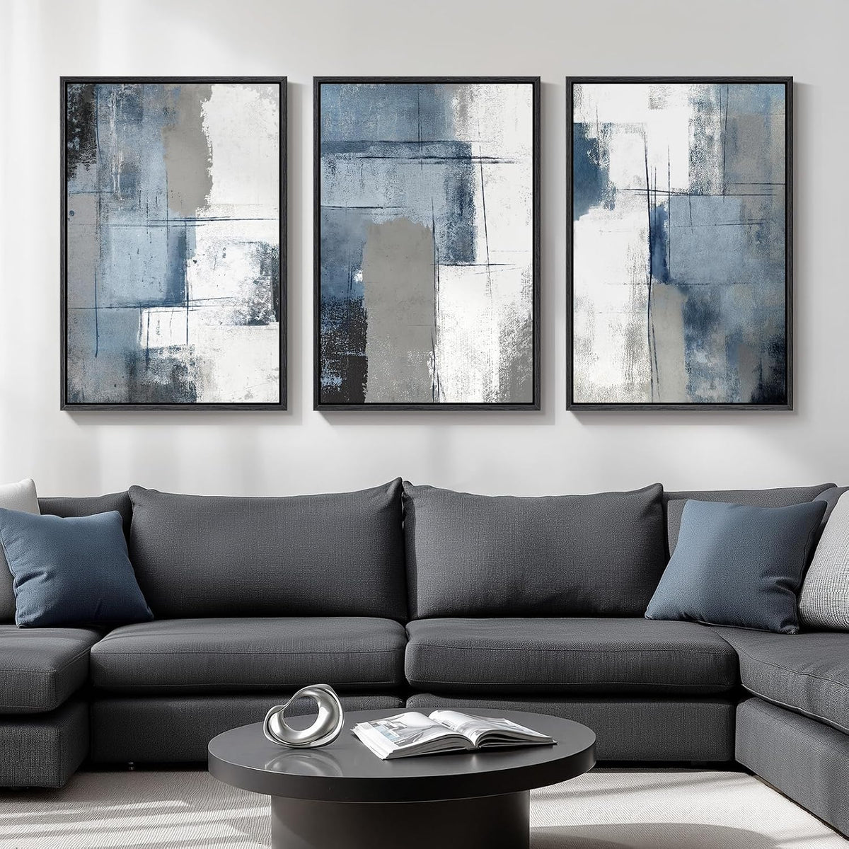 Triptych of blue-gray textured abstract paintings in black frames above a dark gray sofa and round coffee table.