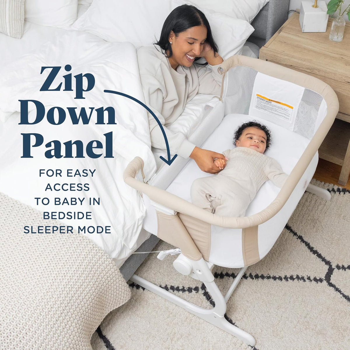 Top-down bedside view of a beige bassinet zipped down beside a bed with a smiling parent and baby inside.