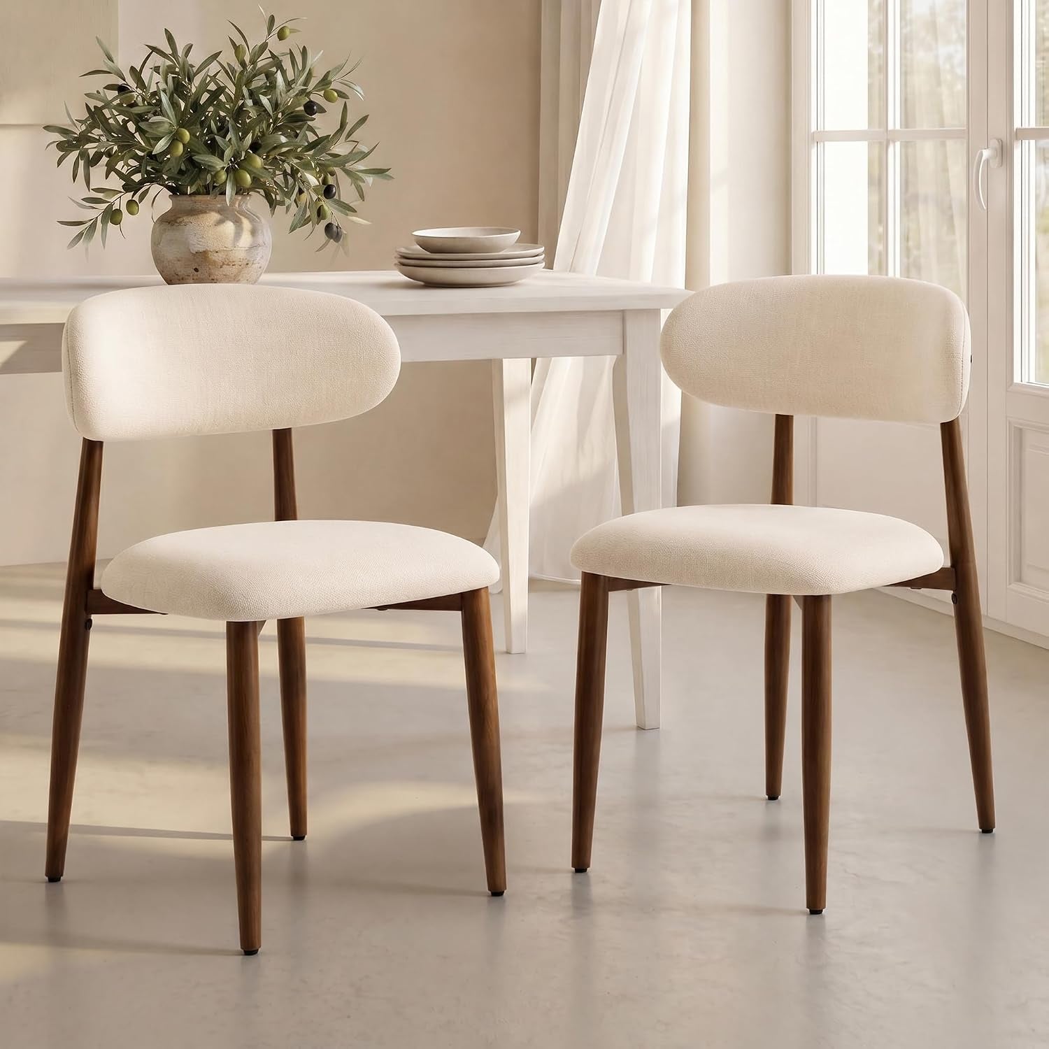 Two beige upholstered dining chairs with warm wood-grain metal legs in front of a pale rectangular table and vase