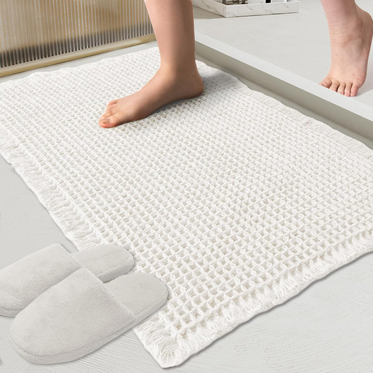Close-up of a white waffle bath mat with a bare foot stepping onto it and slippers nearby.