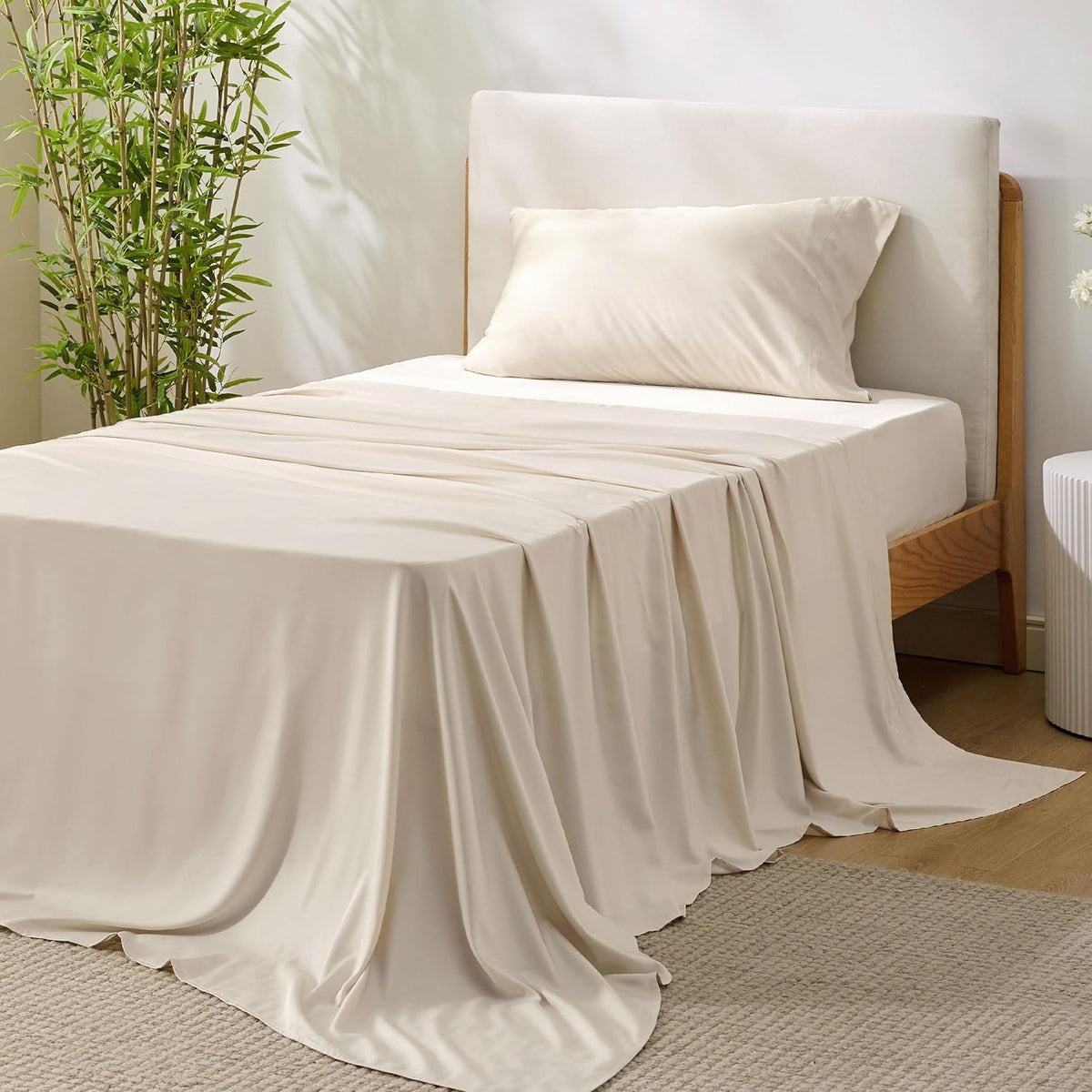 Neutral cream-colored silky sheets flowing over the side of a bed with a light upholstered headboard.