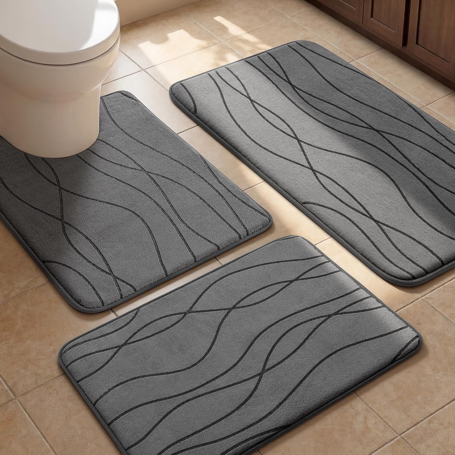 Close view of three dark gray bath mats on tiled floor near toilet and vanity