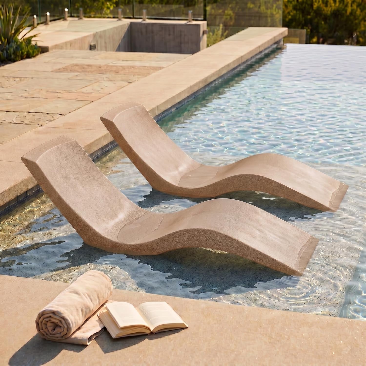 Pair of tan resin loungers set in shallow sun shelf water beside an infinity pool, a rolled towel and open book on the deck.