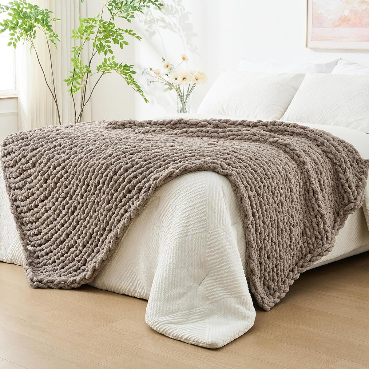 Taupe chunky knit throw spread over the foot of a bed in a bright bedroom with plants and pillows.