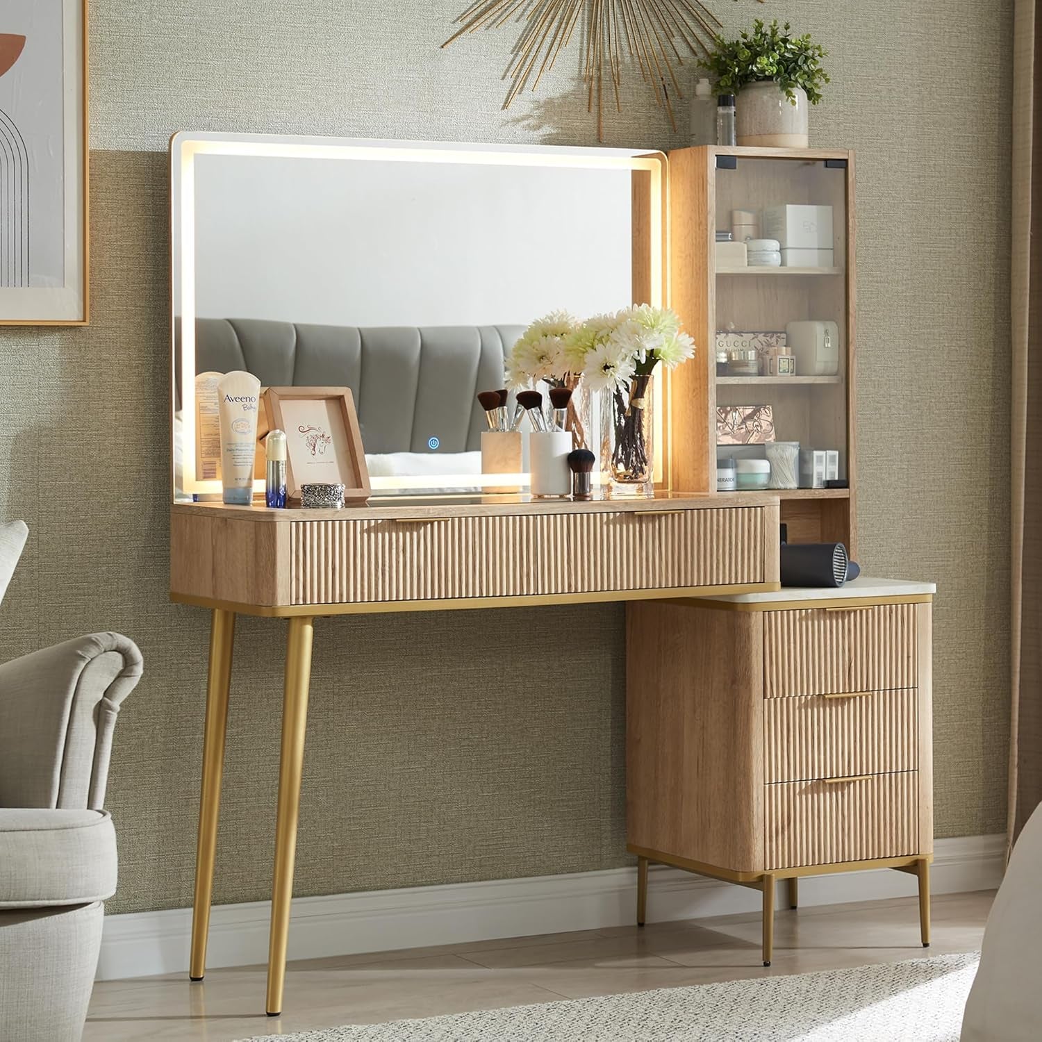 Front view of light wood vanity staged with decor showing glass-front side cabinet, frameless lit mirror and gold tapered leg