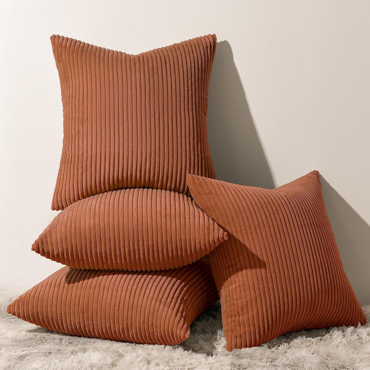 Stack of four ribbed corduroy square pillows in a warm burnt orange shade against a neutral wall.