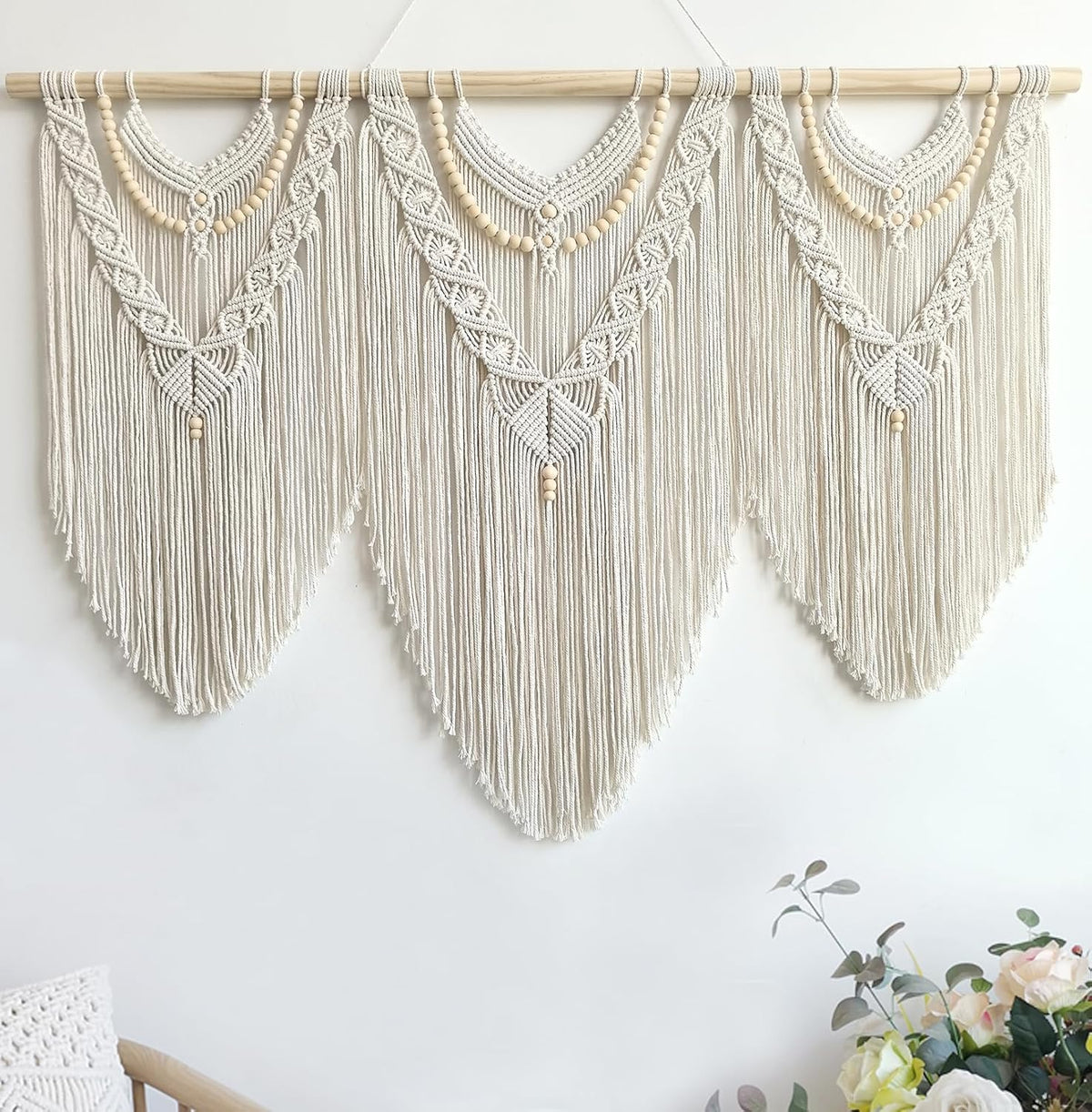 Three-panel macrame filling the upper wall with long V-shaped fringe and a bouquet visible at lower right.