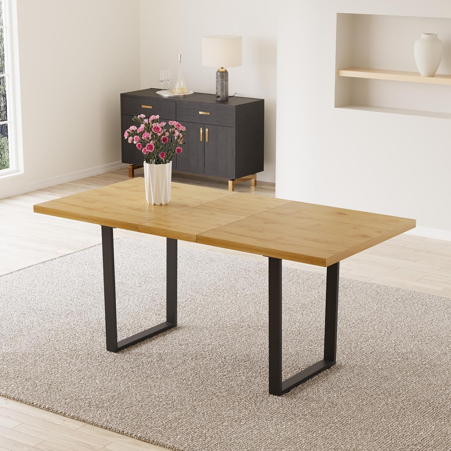 Light wood rectangular table with black metal U-legs on a neutral rug, a white vase with pink flowers centered.
