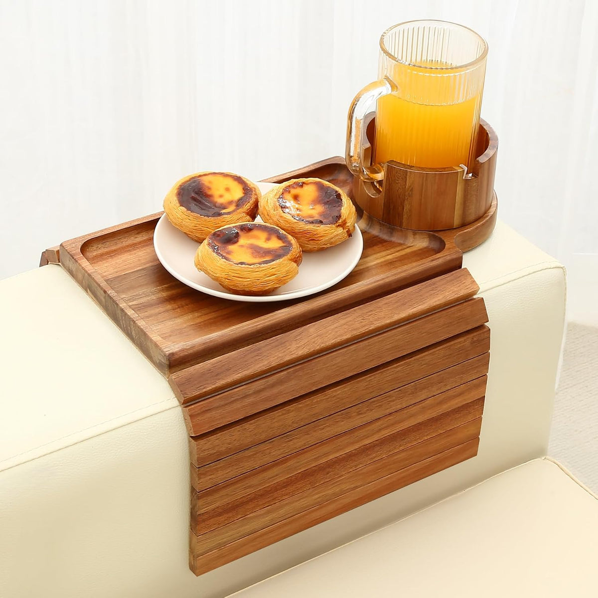 Angled view of a slatted bamboo arm tray draping down a cream sofa arm, holding a plate of three pastries and a mug in the