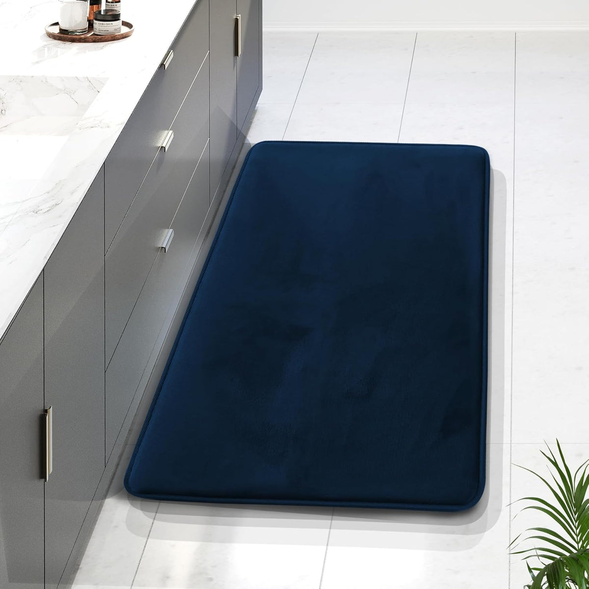 Long navy memory-foam runner placed along a grey kitchen cabinet on white tile floor, partial plant at corner