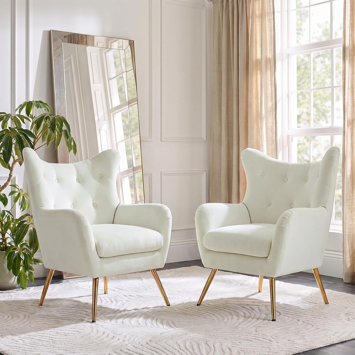 Two off-white wingback chairs in soft light with tall windows and a potted plant nearby.