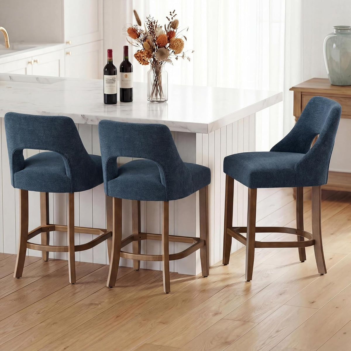 Three navy blue upholstered counter stools with wooden legs tucked at a kitchen island with wine bottles on top