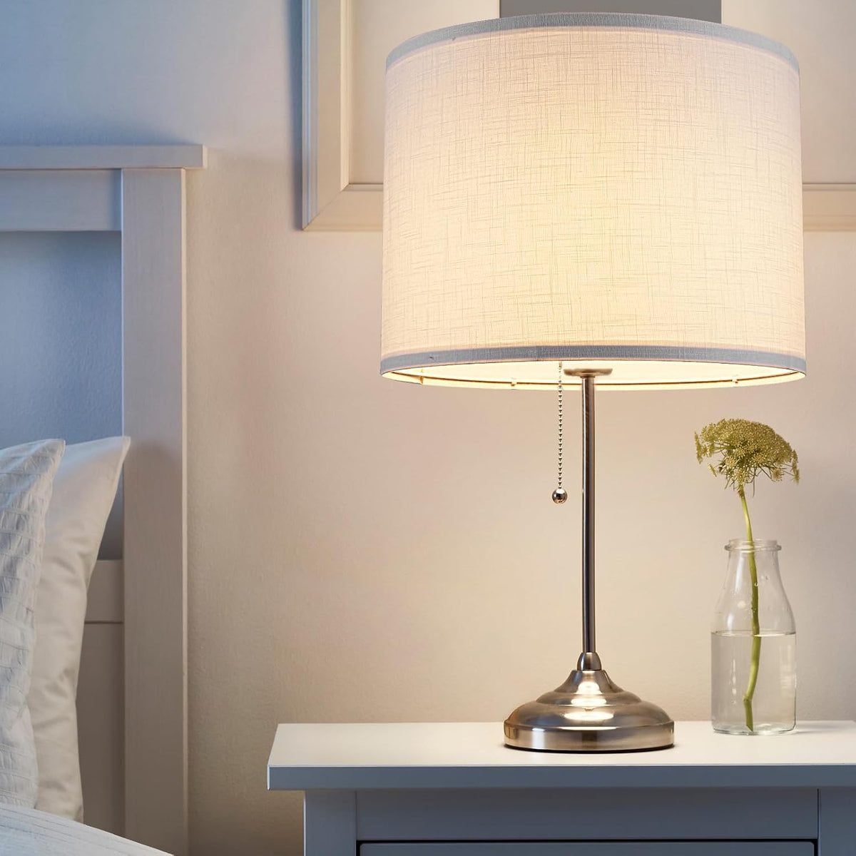 Close-up of a lit white drum shade on a metal bedside lamp with pull-chain, sitting on a nightstand next to a bed.